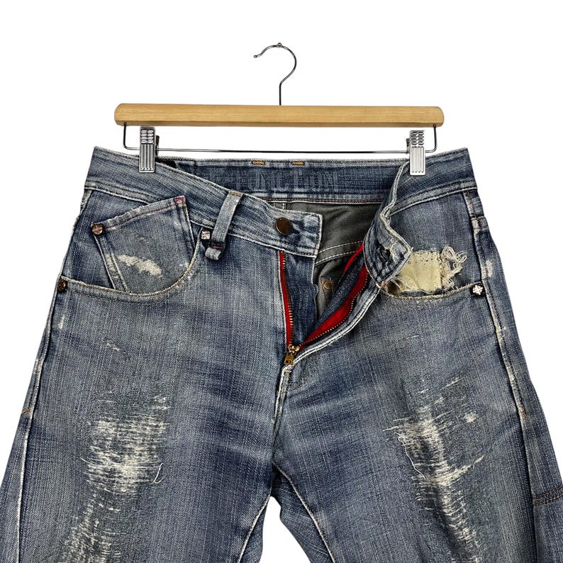 K&ouml;nnte beinhalten: Hellblaue Jeans mit Used-Details, rotem Rei&szlig;verschluss und Knopfverschluss. Die Jeans h&auml;ngen an einem Holzb&uuml;gel. Die Taschen haben einen abgenutzten Look.