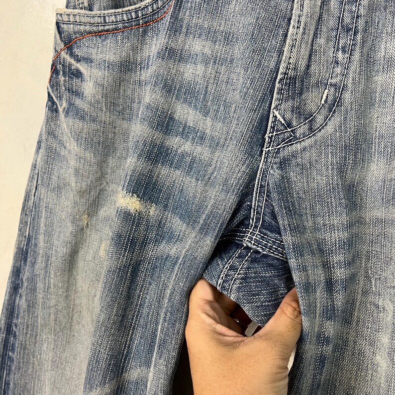 May include: Close-up of light blue denim jeans with distressed details. The jeans have a faded wash, visible wear, and white stitching. The pocket has a red stitch detail.