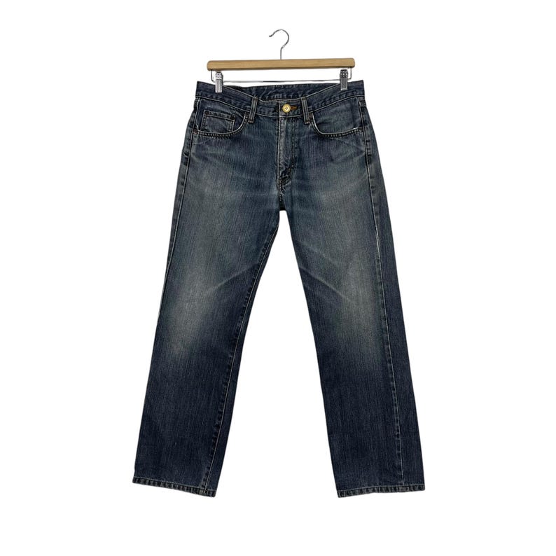May include: Dark wash denim jeans hanging on a wooden hanger. The jeans feature a classic five-pocket design, a button and zip closure, and a straight leg fit. The denim has a faded appearance.