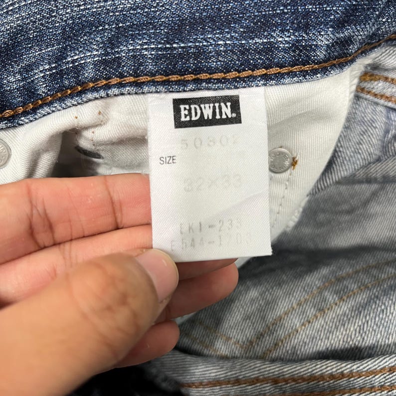 May include: Close-up of a denim jeans label with the brand name "EDWIN" in a black rectangle. The label also shows the size "32x33" and other product codes. The jeans are blue.