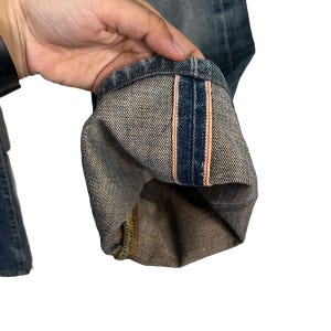 May include: Close-up of the inside of a pair of blue denim jeans with a pink and blue striped seam.