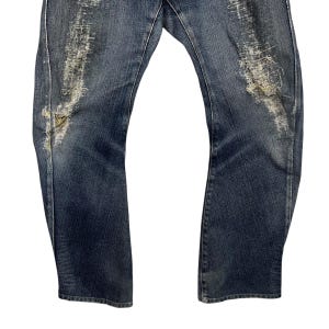 K&ouml;nnte beinhalten: Verblasste blaue Jeans mit Distressed-Details. Die Jeans haben gerissene Bereiche mit freiliegenden F&auml;den und ein abgenutztes Aussehen. Der Denim hat eine mittlere Waschung mit helleren Bereichen durch Abnutzung. Die Jeans haben ein klassisches F&uuml;nf-Taschen-Design.
