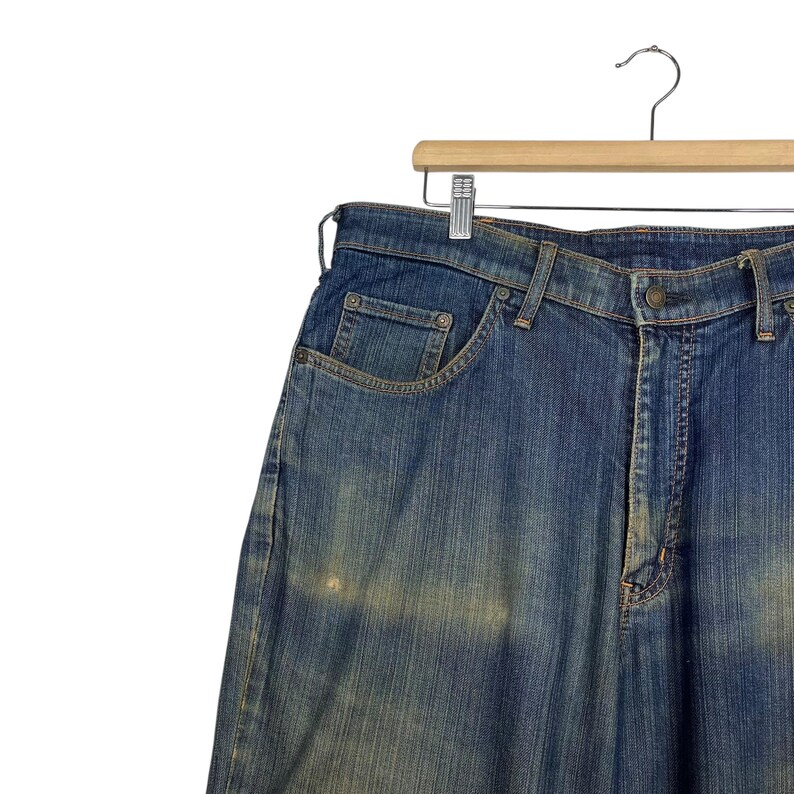 K&ouml;nnte beinhalten: Blaue Jeans aus Denim, die an einem Holzb&uuml;gel h&auml;ngen. Die Jeans haben ein klassisches F&uuml;nf-Taschen-Design und eine verwaschene Optik. Der Denim ist mittelblau mit helleren Bereichen, was ihm einen Used-Look verleiht. Die Jeans werden vor wei&szlig;em Hintergrund pr&auml;sentiert.