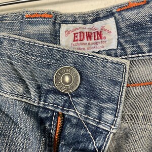 May include: Close-up of blue denim jeans with orange stitching and a silver button. The jeans feature a label that reads "EDWIN" and "Exclusive design for lasting good look."
