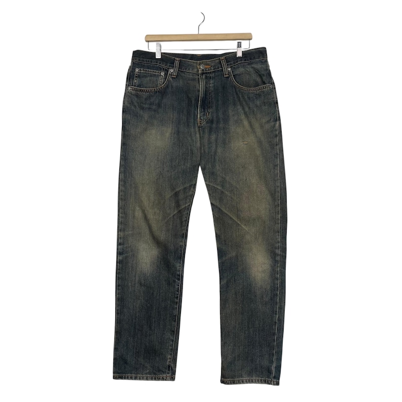 May include: Blue denim jeans with a faded wash and a classic five-pocket design. The jeans are hanging on a wooden hanger against a white background. The denim has a worn appearance.