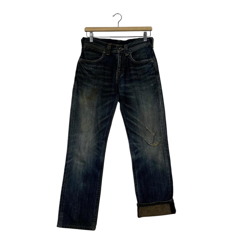 May include: Dark wash denim jeans with a distressed look, featuring a knee rip and rolled-up cuffs. The jeans are hanging on a wooden hanger against a white background. The jeans appear to be a straight-leg style.