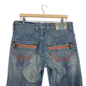 May include: Light blue denim jeans with orange zipper pockets and stitching. The jeans have a classic five-pocket design with a branded patch on the back. The jeans are hanging on a wooden hanger.
