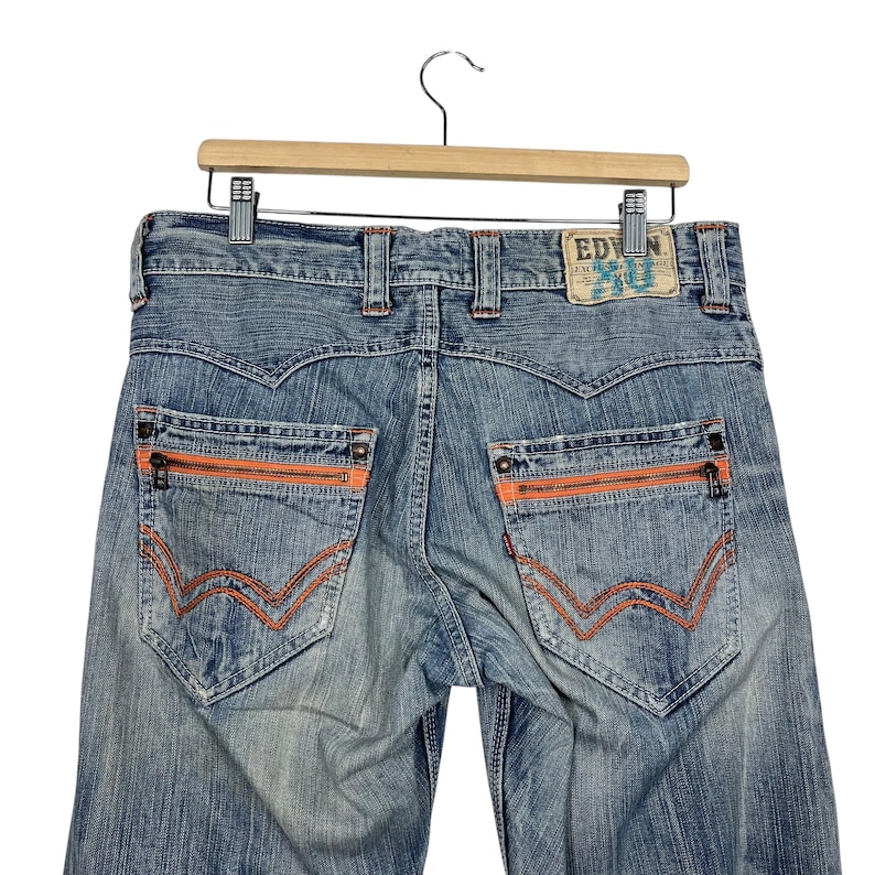 May include: Light blue denim jeans with orange zipper pockets and stitching. The jeans have a classic five-pocket design with a branded patch on the back. The jeans are hanging on a wooden hanger.