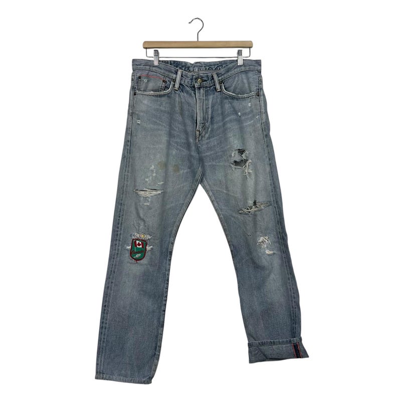 May include: Light blue denim jeans with distressed details, including rips and patches. The jeans have a classic five-pocket design and are cuffed at the ankles. A colorful patch is visible on the left leg. The jeans are hanging on a wooden hanger.