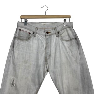 May include: Light gray denim jeans with a distressed knee detail, hanging on a wooden hanger. The jeans have classic five-pocket styling with copper rivets and a button closure. The denim appears faded and worn.