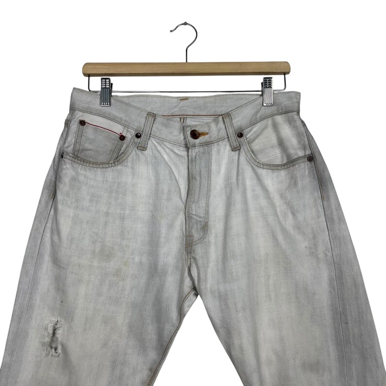 May include: Light gray denim jeans with a distressed knee detail, hanging on a wooden hanger. The jeans have classic five-pocket styling with copper rivets and a button closure. The denim appears faded and worn.