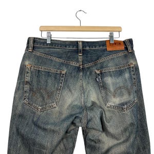 May include: Faded blue denim jeans with a classic five-pocket design. The jeans are displayed on a wooden hanger, showcasing the back pockets and the brand's leather patch. The denim has a worn, vintage look.