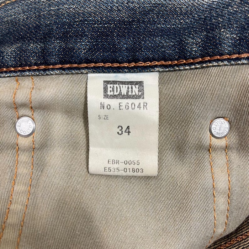 May include: Close-up of the inside of a pair of blue denim jeans. A white tag displays the brand "EDWIN", the model number "E604R", and the size "34". Two silver metal buttons are visible.