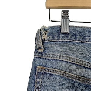 K&ouml;nnte beinhalten: Nahaufnahme einer blauen Jeans, die an einem Holzkleiderb&uuml;gel h&auml;ngt. Die Jeans hat einen abgenutzten, getragenen Look mit einem ausgefransten Rand an der Taille. Die Ges&auml;&szlig;tasche ist sichtbar, und die Jeans ist mit einer Metallklammer am B&uuml;gel befestigt.