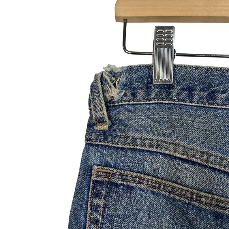 K&ouml;nnte beinhalten: Nahaufnahme einer blauen Jeans, die an einem Holzkleiderb&uuml;gel h&auml;ngt. Die Jeans hat einen abgenutzten, getragenen Look mit einem ausgefransten Rand an der Taille. Die Ges&auml;&szlig;tasche ist sichtbar, und die Jeans ist mit einer Metallklammer am B&uuml;gel befestigt.