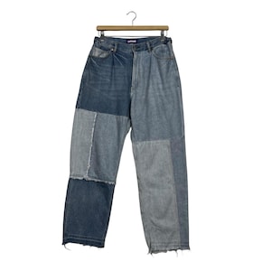 May include: Patchwork denim jeans in shades of blue and gray hang from a wooden hanger. The jeans feature a unique design with various denim patches sewn together. The raw hem adds a casual touch to the garment. The jeans are a stylish addition to any wardrobe.
