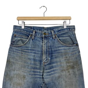 K&ouml;nnte beinhalten: Nahaufnahme einer blauen Jeans, die an einem Holzb&uuml;gel h&auml;ngt. Die Jeans weist Gebrauchsspuren auf, mit verblichenen Stellen und Verf&auml;rbungen. Die Jeans hat ein klassisches F&uuml;nf-Taschen-Design und einen Knopf- und Rei&szlig;verschluss.