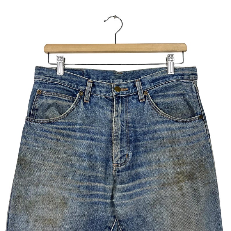 K&ouml;nnte beinhalten: Nahaufnahme einer blauen Jeans, die an einem Holzb&uuml;gel h&auml;ngt. Die Jeans weist Gebrauchsspuren auf, mit verblichenen Stellen und Verf&auml;rbungen. Die Jeans hat ein klassisches F&uuml;nf-Taschen-Design und einen Knopf- und Rei&szlig;verschluss.