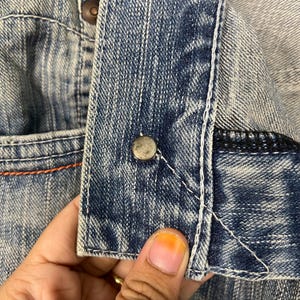 May include: Close-up of a denim jacket detail. The image shows a section of the jacket with a button, white stitching, and a pocket. The denim is a faded blue color, with visible texture and wear.