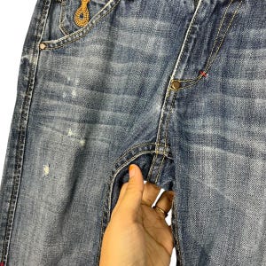 K&ouml;nnte beinhalten: Nahaufnahme einer blauen Jeans mit Used-Details. Die Jeans hat einen Knopfverschluss, sichtbare N&auml;hte und eine kleine goldfarbene Verzierung in der N&auml;he der Tasche. Eine Hand greift in die Tasche.