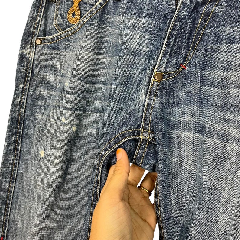 K&ouml;nnte beinhalten: Nahaufnahme einer blauen Jeans mit Used-Details. Die Jeans hat einen Knopfverschluss, sichtbare N&auml;hte und eine kleine goldfarbene Verzierung in der N&auml;he der Tasche. Eine Hand greift in die Tasche.