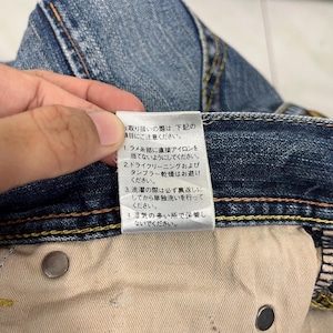 May include: Close-up of blue denim jeans with a white tag. The tag has Japanese text with care instructions. The jeans have yellow stitching and metal rivets. The fabric is faded and worn.