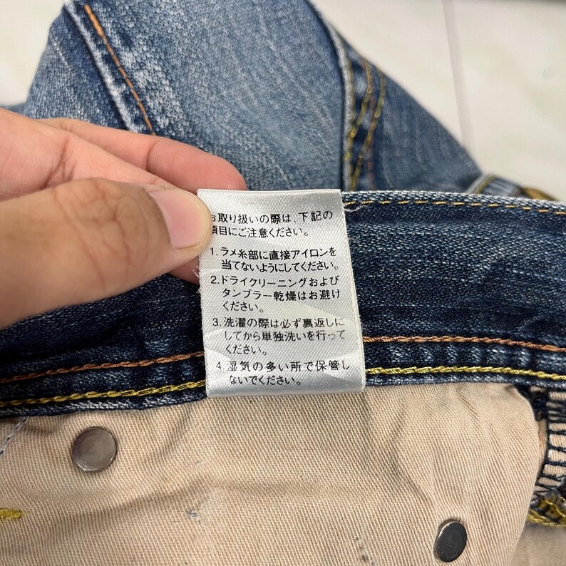 May include: Close-up of blue denim jeans with a white tag. The tag has Japanese text with care instructions. The jeans have yellow stitching and metal rivets. The fabric is faded and worn.