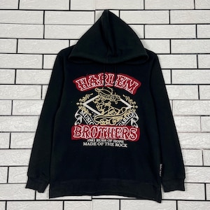 Może przedstawiać: Czarna bluza z kapturem z grafiką z napisem "HARLEM BROTHERS MUSIC GARAGE" w czerwono-złotych literach. Grafika przedstawia postać z kreskówki oraz tekst "1981 RUSH OF HOPE MADE OF THE ROCK".