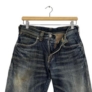 May include: Close-up of a pair of blue denim jeans hanging on a wooden hanger. The jeans have a classic five-pocket design, a button and zipper closure, and show signs of wear and fading, giving them a vintage look.