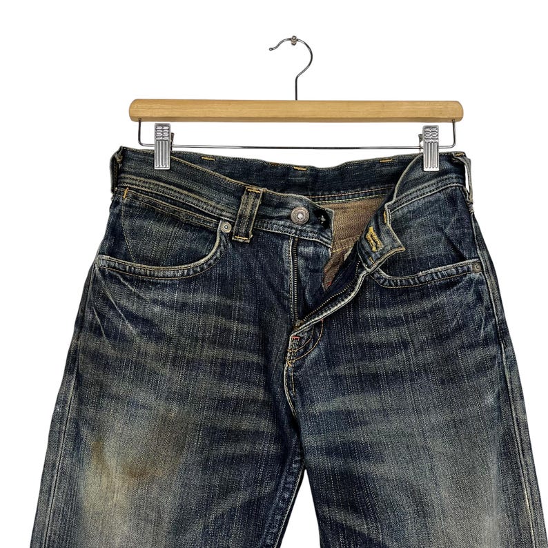 May include: Close-up of a pair of blue denim jeans hanging on a wooden hanger. The jeans have a classic five-pocket design, a button and zipper closure, and show signs of wear and fading, giving them a vintage look.