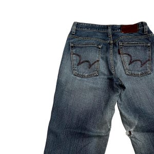 Op de afbeelding: Verbleekte blauwe denim jeans met een versleten uiterlijk. Voorzien van twee achterzakken met een kenmerkend gestikt ontwerp, een bruin leren label en een klein rood label. De jeans vertonen tekenen van slijtage, waaronder een gat bij de knie.
