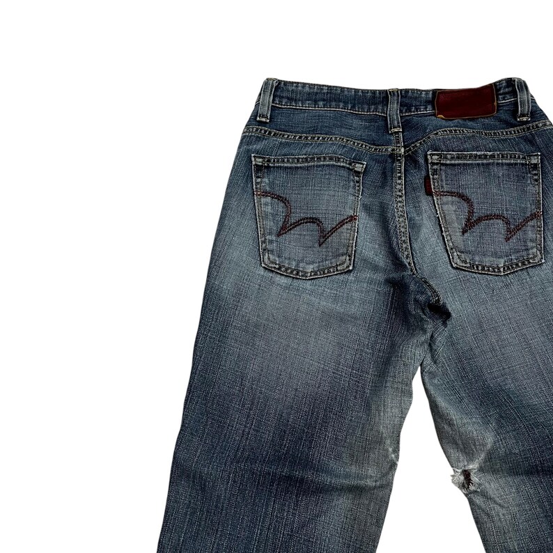Op de afbeelding: Verbleekte blauwe denim jeans met een versleten uiterlijk. Voorzien van twee achterzakken met een kenmerkend gestikt ontwerp, een bruin leren label en een klein rood label. De jeans vertonen tekenen van slijtage, waaronder een gat bij de knie.