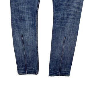 K&ouml;nnte beinhalten: Nahaufnahme einer blauen Jeans mit vertikalen Schlitzdetails am unteren Saum jedes Beins. Die Jeans hat ein klassisches F&uuml;nf-Taschen-Design und eine verblasste Waschung, die Gebrauchsspuren aufweist.