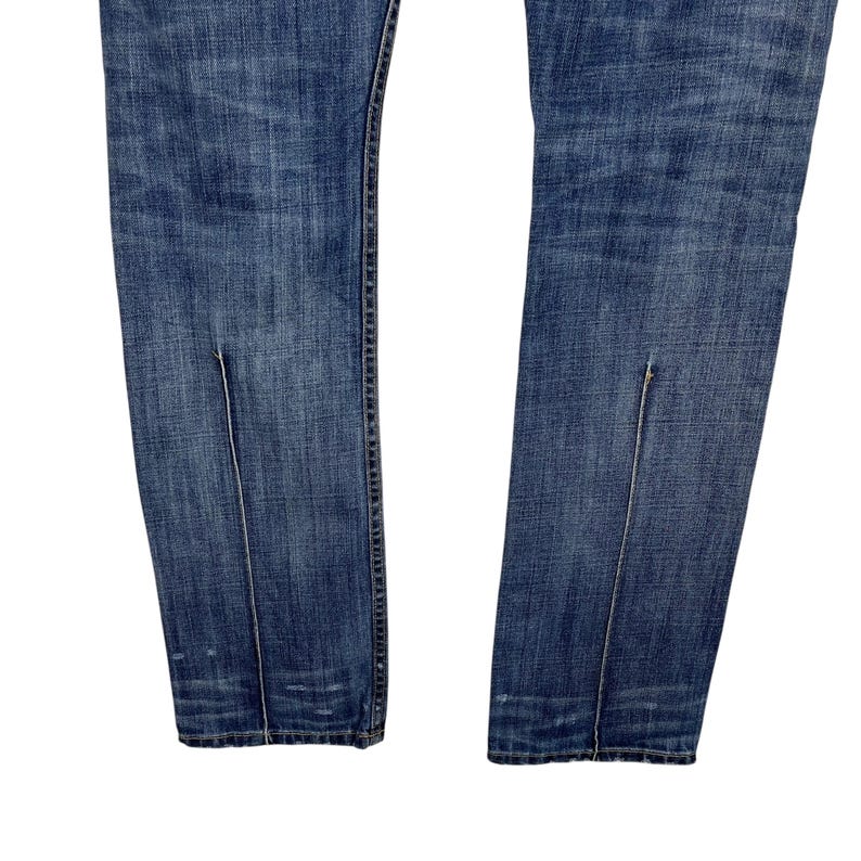 K&ouml;nnte beinhalten: Nahaufnahme einer blauen Jeans mit vertikalen Schlitzdetails am unteren Saum jedes Beins. Die Jeans hat ein klassisches F&uuml;nf-Taschen-Design und eine verblasste Waschung, die Gebrauchsspuren aufweist.