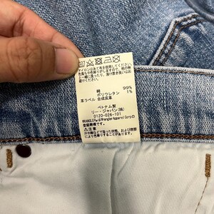 K&ouml;nnte beinhalten: Nahaufnahme eines Jeansetiketts mit japanischem Text und Pflegesymbolen. Das Etikett enth&auml;lt Angaben zur Stoffzusammensetzung: 99 % Baumwolle, 1 % Polyurethan. Die Marke ist Wrangler, hergestellt in Vietnam.