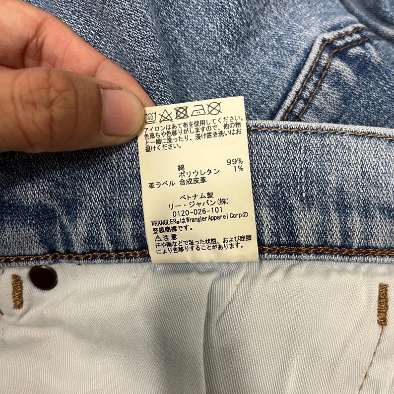 K&ouml;nnte beinhalten: Nahaufnahme eines Jeansetiketts mit japanischem Text und Pflegesymbolen. Das Etikett enth&auml;lt Angaben zur Stoffzusammensetzung: 99 % Baumwolle, 1 % Polyurethan. Die Marke ist Wrangler, hergestellt in Vietnam.