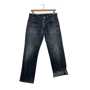May include: Dark wash denim jeans hanging on a wooden hanger. The jeans feature a classic five-pocket design, a button and zip closure, and a folded cuff at the hem. The denim has a faded appearance.