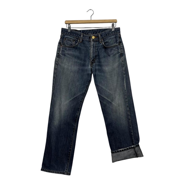 May include: Dark wash denim jeans hanging on a wooden hanger. The jeans feature a classic five-pocket design, a button and zip closure, and a folded cuff at the hem. The denim has a faded appearance.