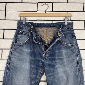 May include: Blue denim jeans with a button and zipper closure, hanging on a wooden hanger. The jeans have a faded wash and visible stitching details. The pockets and waistband are clearly visible.