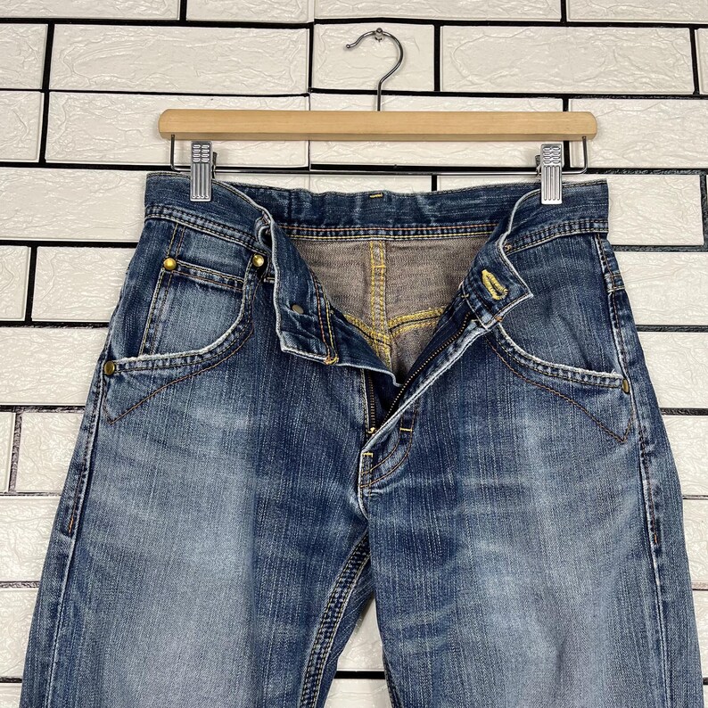 May include: Blue denim jeans with a button and zipper closure, hanging on a wooden hanger. The jeans have a faded wash and visible stitching details. The pockets and waistband are clearly visible.
