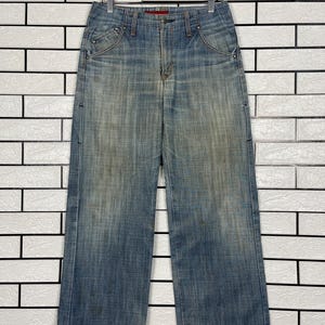 May include: Faded blue denim jeans with a wide leg design. The jeans are hanging on a wooden hanger against a white brick wall. The jeans have a button and zipper closure, with pockets on the front and back. The denim has a worn, distressed look.
