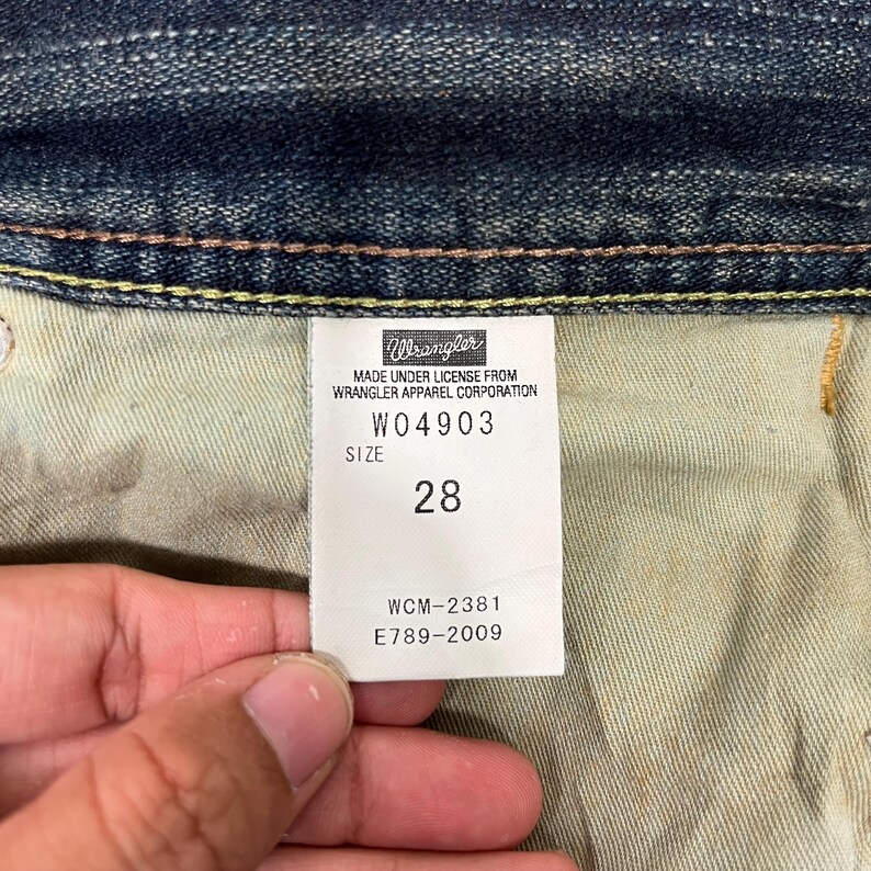 May include: Close-up of a Wrangler jeans tag. The tag reads "MADE UNDER LICENSE FROM WRANGLER APPAREL CORPORATION" with the style number W04903 and size 28. Additional codes WCM-2381 and E789-2009 are also visible.