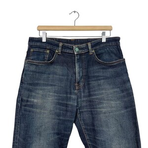 Puede incluir: Vaqueros de mezclilla azul oscuro colgados de una percha de madera. Los vaqueros tienen un dise&ntilde;o cl&aacute;sico de cinco bolsillos con cierre de bot&oacute;n y cremallera. La mezclilla tiene un aspecto descolorido, con sutiles marcas de desgaste.