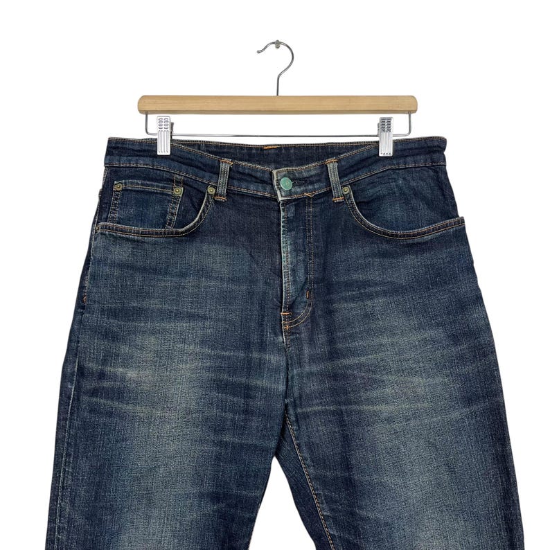 Puede incluir: Vaqueros de mezclilla azul oscuro colgados de una percha de madera. Los vaqueros tienen un dise&ntilde;o cl&aacute;sico de cinco bolsillos con cierre de bot&oacute;n y cremallera. La mezclilla tiene un aspecto descolorido, con sutiles marcas de desgaste.