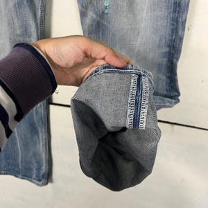 May include: Close-up of light blue denim jeans with a distressed look. The jeans have a folded cuff, revealing the interior fabric and stitching. The cuff is held up by a person's hand, showcasing the details of the denim.