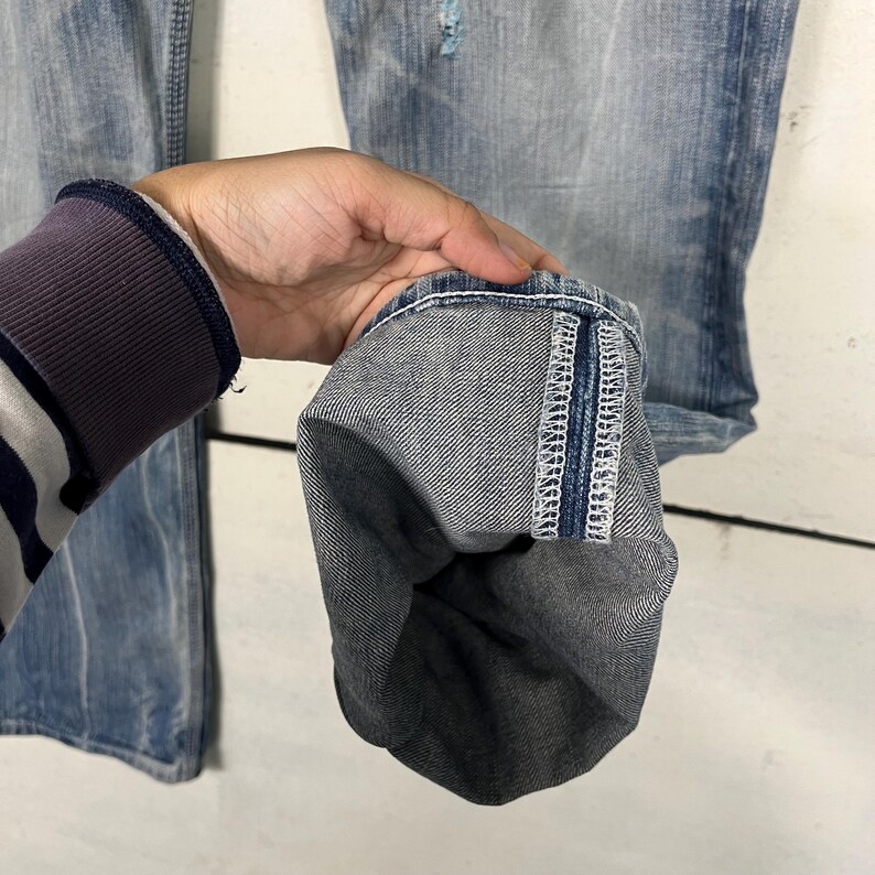 May include: Close-up of light blue denim jeans with a distressed look. The jeans have a folded cuff, revealing the interior fabric and stitching. The cuff is held up by a person's hand, showcasing the details of the denim.
