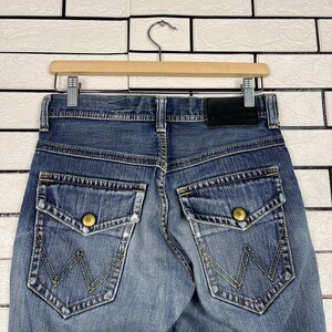 May include: Close-up of the back of blue denim jeans with two large pockets featuring gold button closures and a yellow stitched "W" design. The jeans are hanging on a wooden hanger against a white brick wall.
