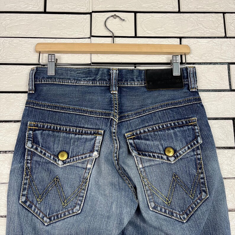 May include: Close-up of the back of blue denim jeans with two large pockets featuring gold button closures and a yellow stitched "W" design. The jeans are hanging on a wooden hanger against a white brick wall.
