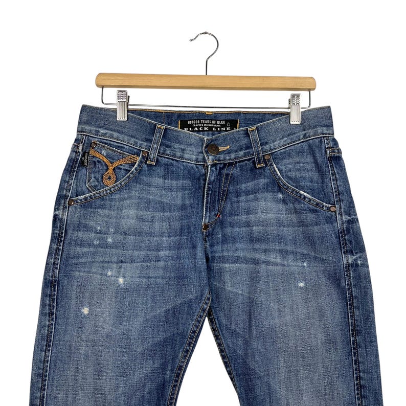 K&ouml;nnte beinhalten: Blaue Jeans aus Denim mit einem Used-Look, mit dekorativen goldfarbenen Ziern&auml;hten an der Vordertasche. Die Jeans h&auml;ngen an einem Holzb&uuml;gel. Der Text "HIGHER TEARS OF BLUE BLACK LINE" ist auf dem Bund zu sehen.