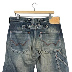 May include: Faded blue denim jeans with red stitching on the back pockets and a leather patch. The jeans have a belt loop detail and are suspended from a wooden hanger. The jeans are a classic style.